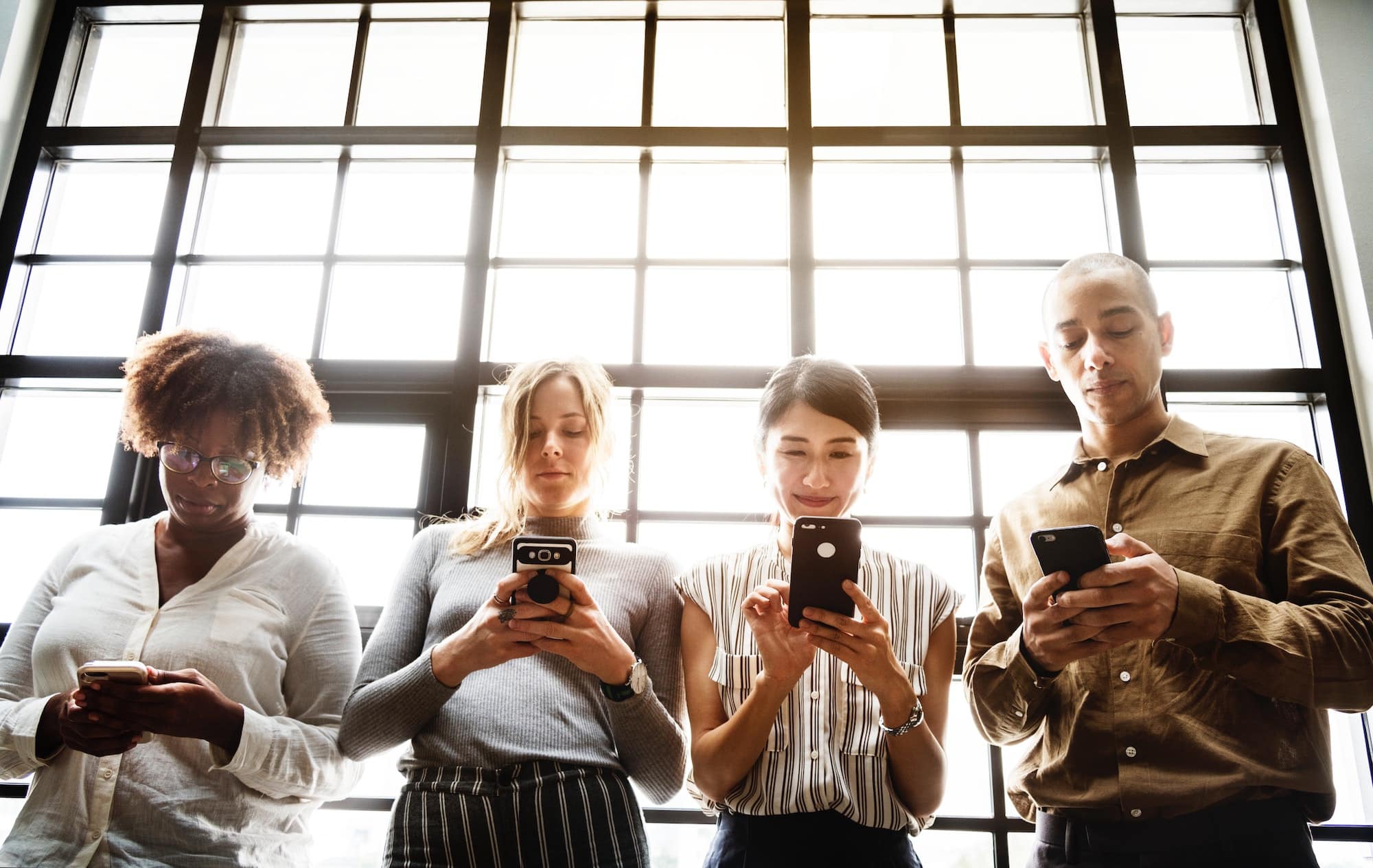 Four people browse smartphones backlit by window panes